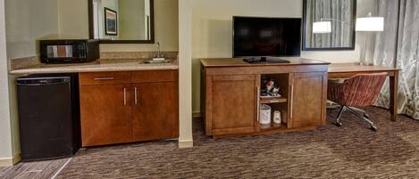 A hotel room with a kitchenette, a TV, a desk with a chair, and a large window with curtains.