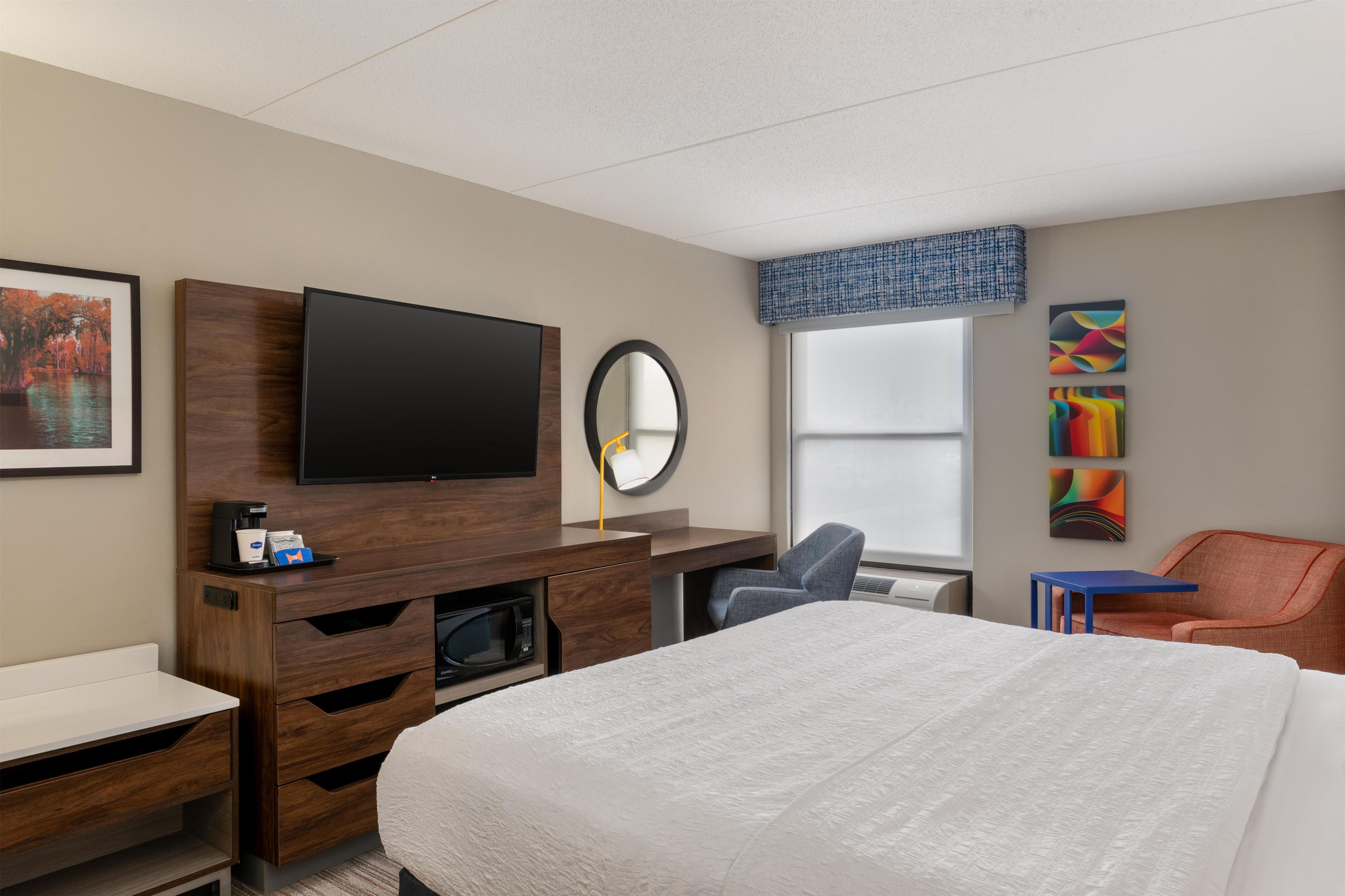 A hotel room with a bed, a TV, a desk, a chair, a microwave, and artwork on the wall.