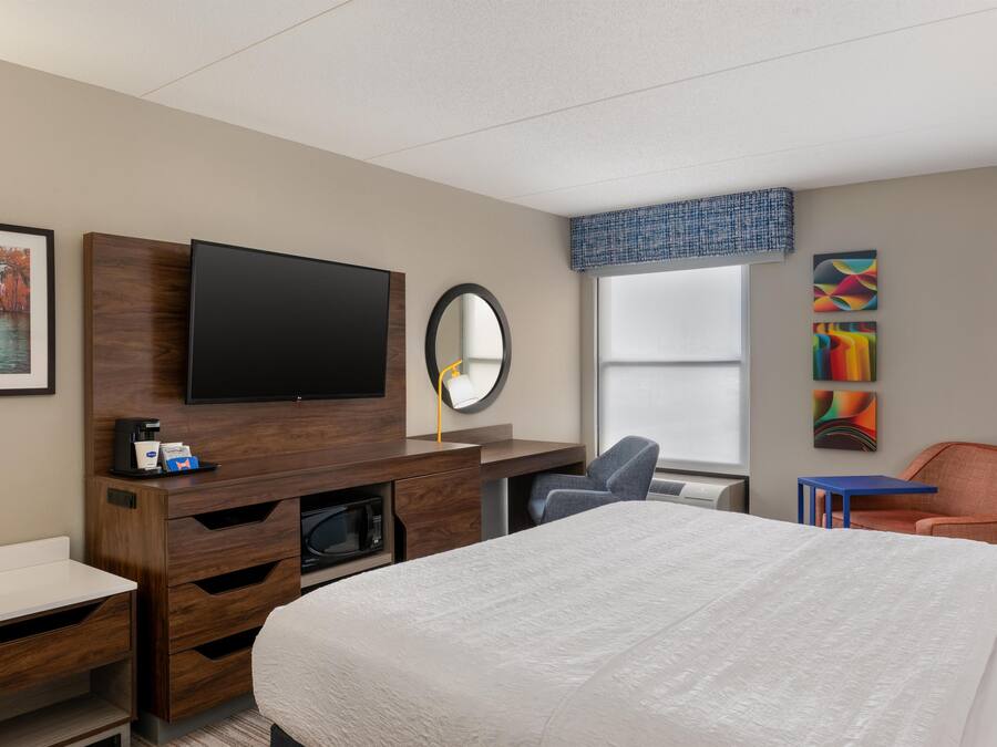 A hotel room with a bed, a TV, a desk, a chair, a microwave, and artwork on the wall.