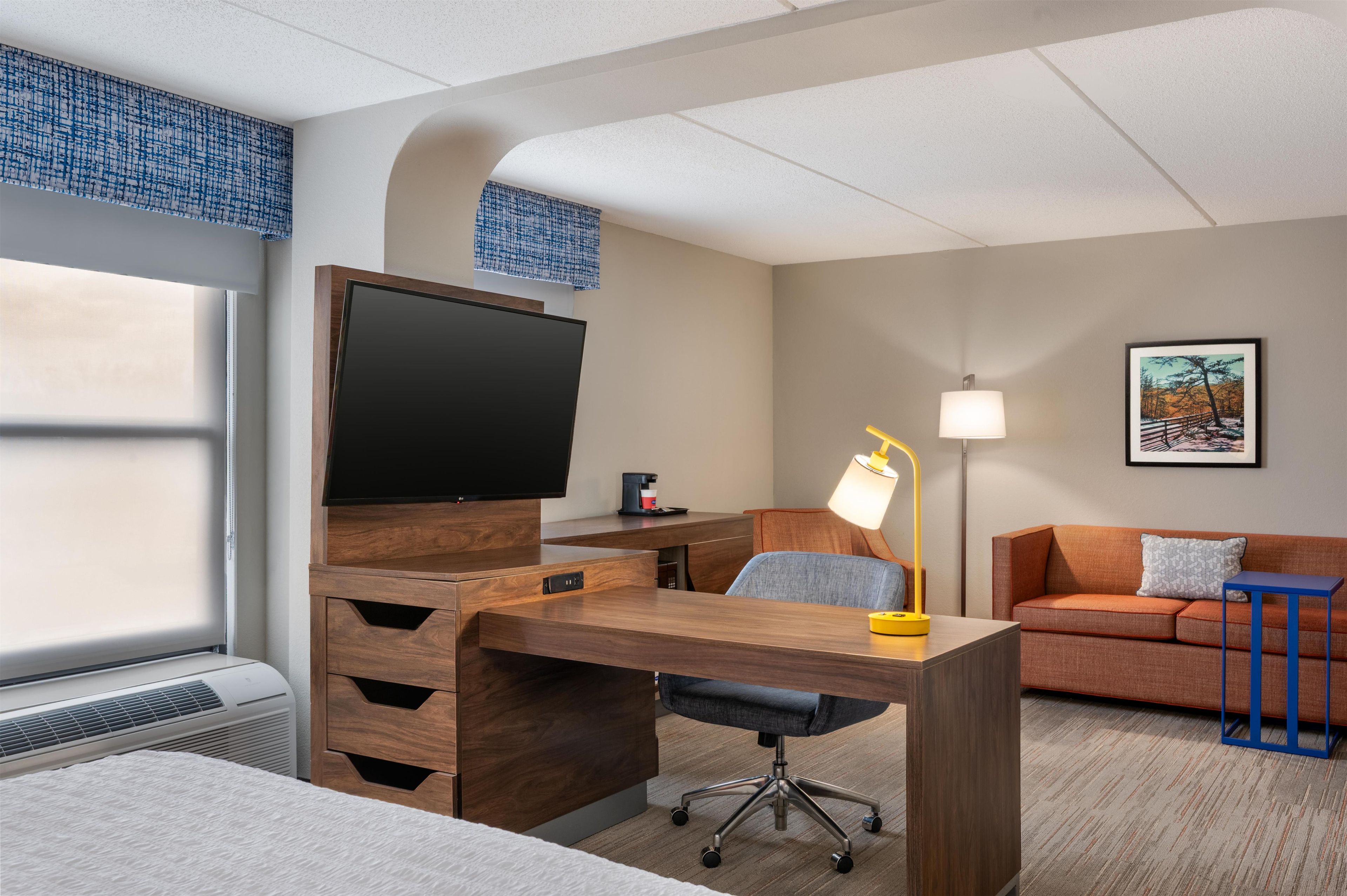 A hotel room with a wooden desk, a flat-screen TV, a desk lamp, a chair, a sofa, a coffee maker, and a framed picture on the wall.