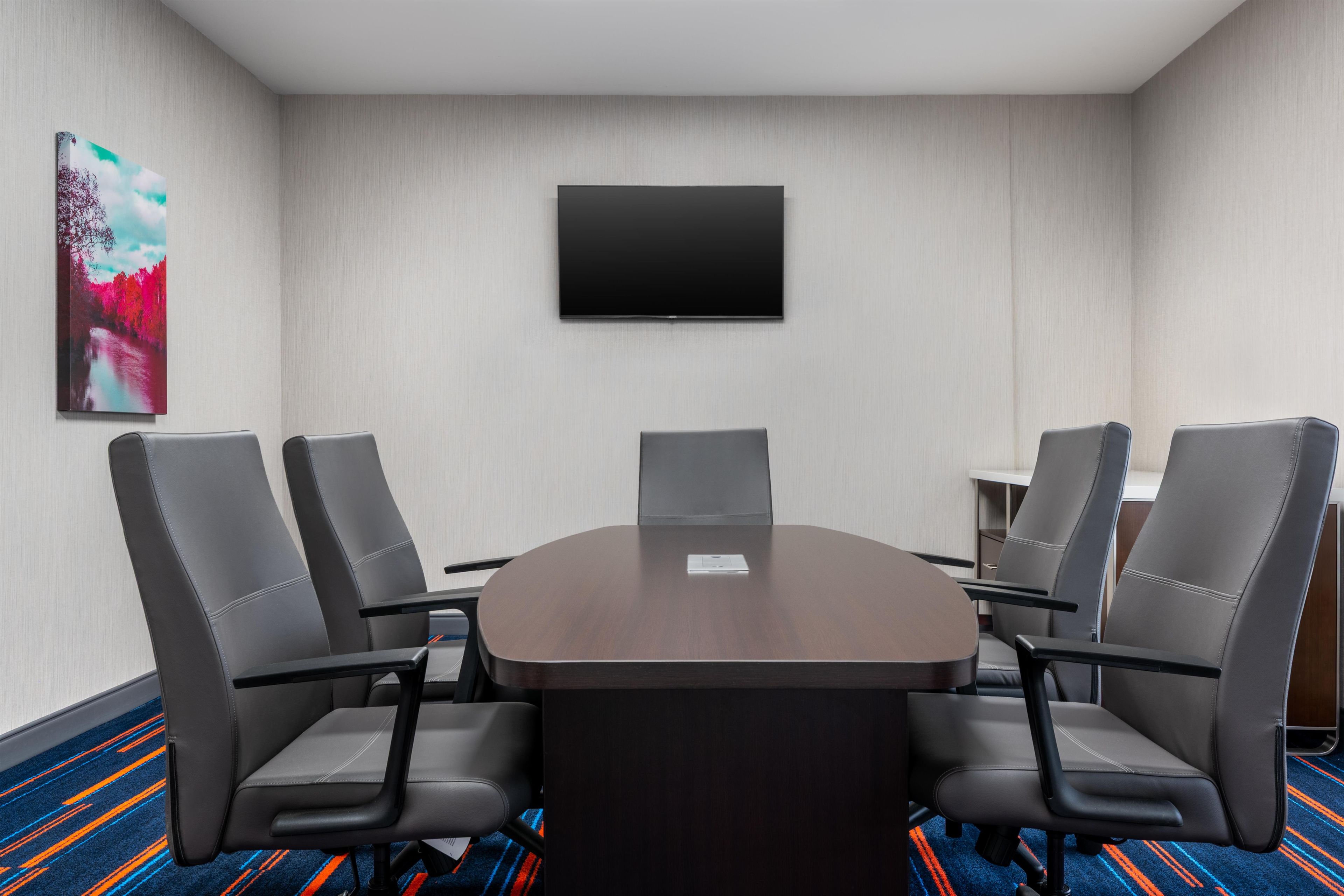 A conference room with a round table, six chairs, a flat-screen TV on the wall, and a colorful carpet.