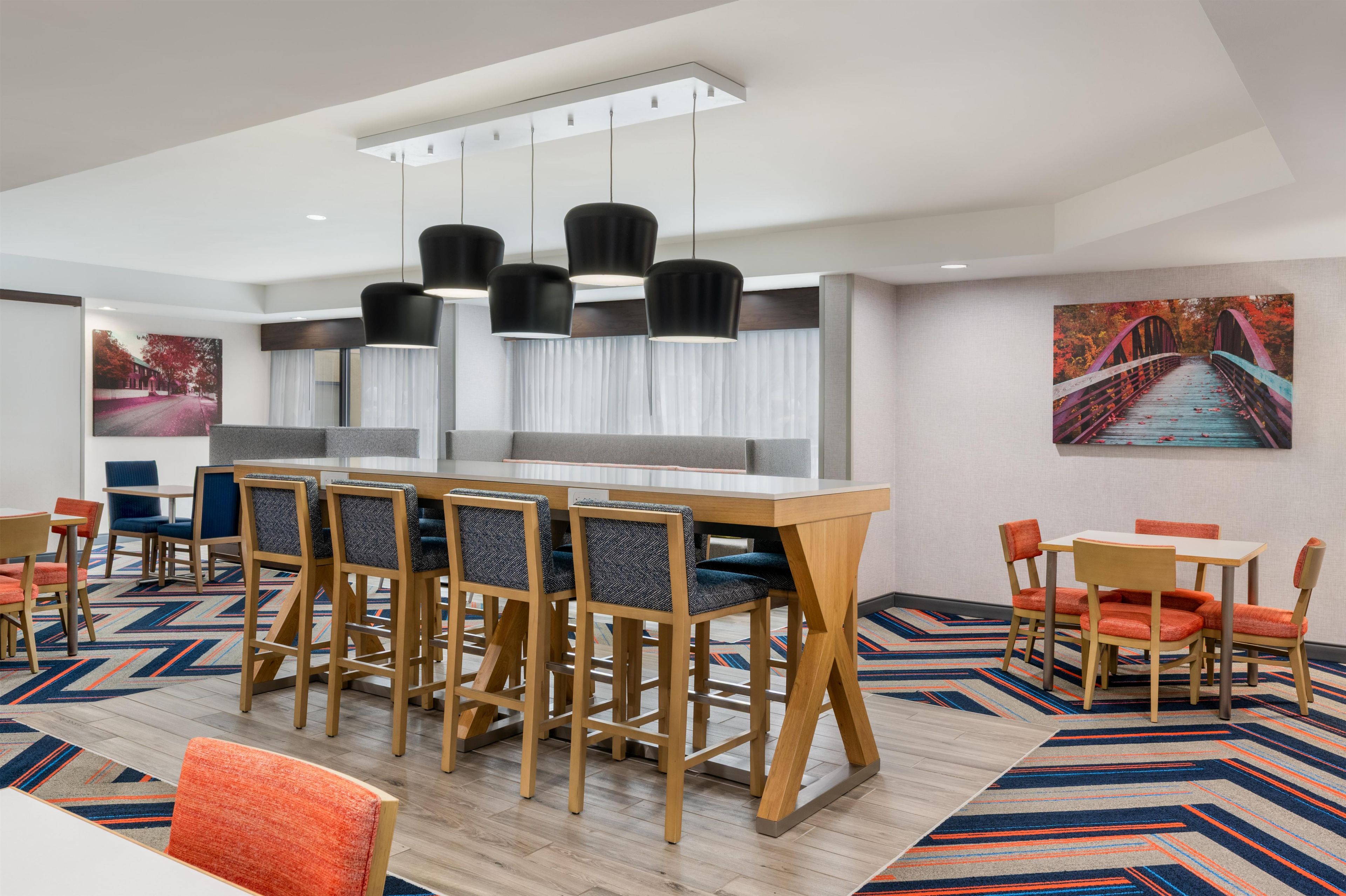 A modern hotel room with a large dining table, multiple chairs, and a view of a bridge.