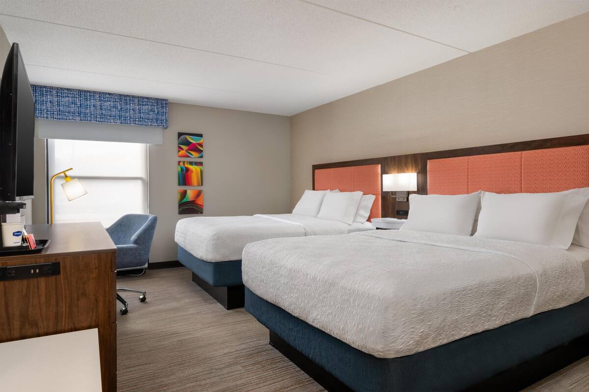 A hotel room with two beds, a desk, a chair, a TV, and a window with a view.