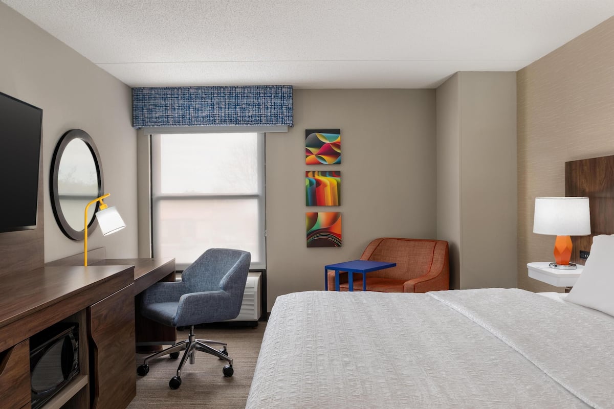 A hotel room with a bed, desk, chairs, a TV, a window, and artwork on the wall.