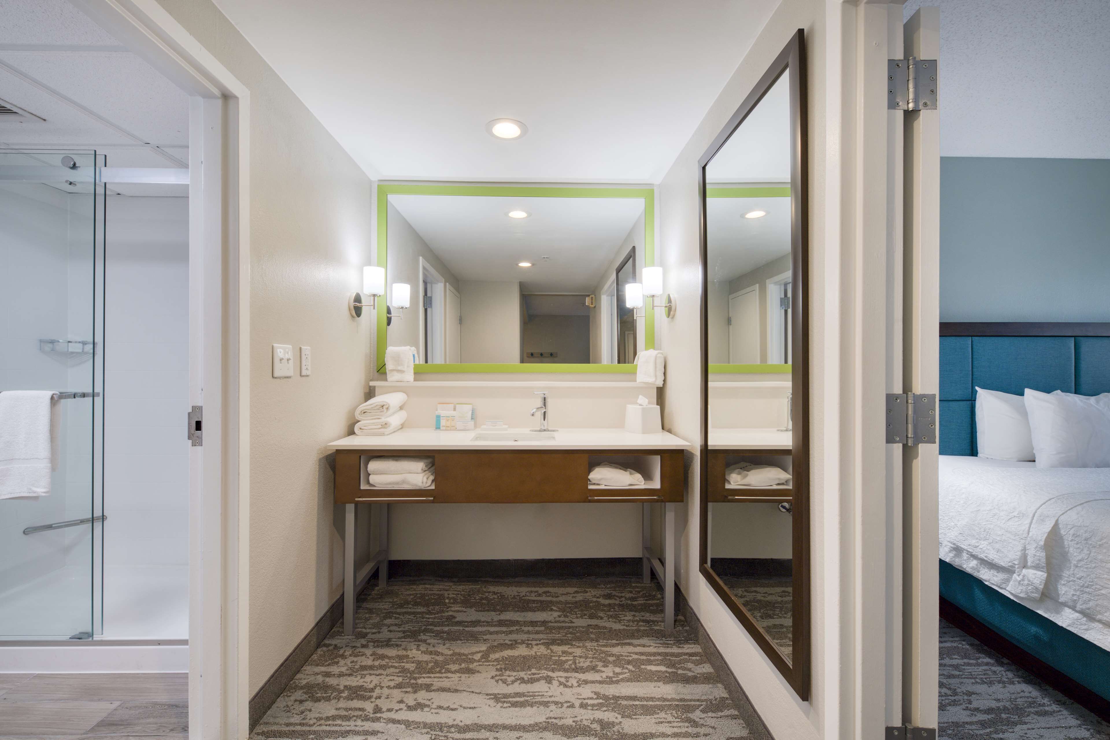 A hotel bathroom with a glass shower, a sink with a mirror, and a bed with white bedding.