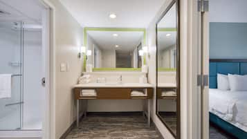A hotel bathroom with a glass shower, a sink with a mirror, and a bed with white bedding.