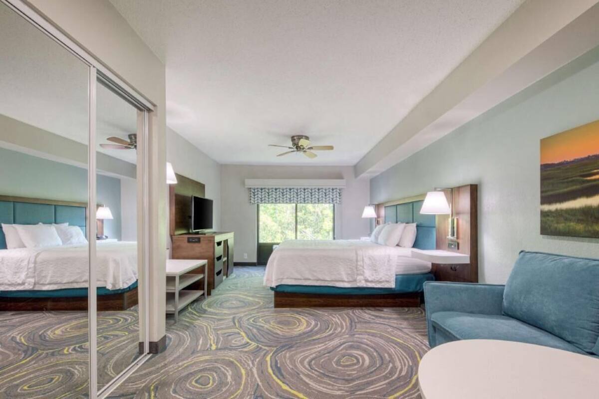 A hotel room with two beds, a desk, a chair, a sofa, a ceiling fan, and a window with a view.