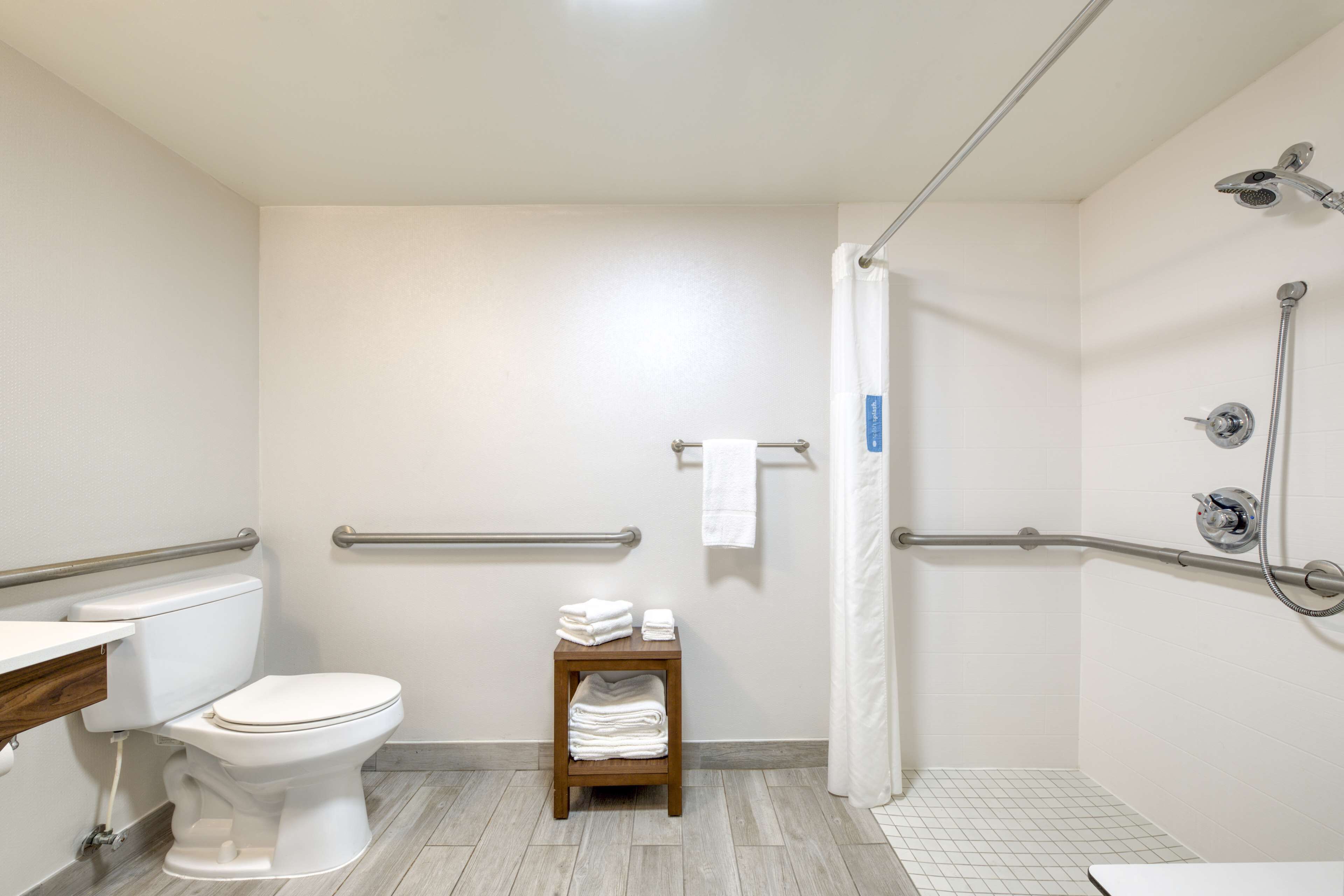 Une salle de bain comprenant des toilettes, un lavabo, une douche équipée de barres d’appui et une petite étagère avec des serviettes.