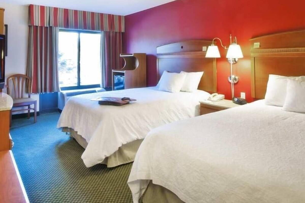 Habitación de hotel con dos camas, un escritorio, una silla, un ventanal con cortinas y una lámpara.