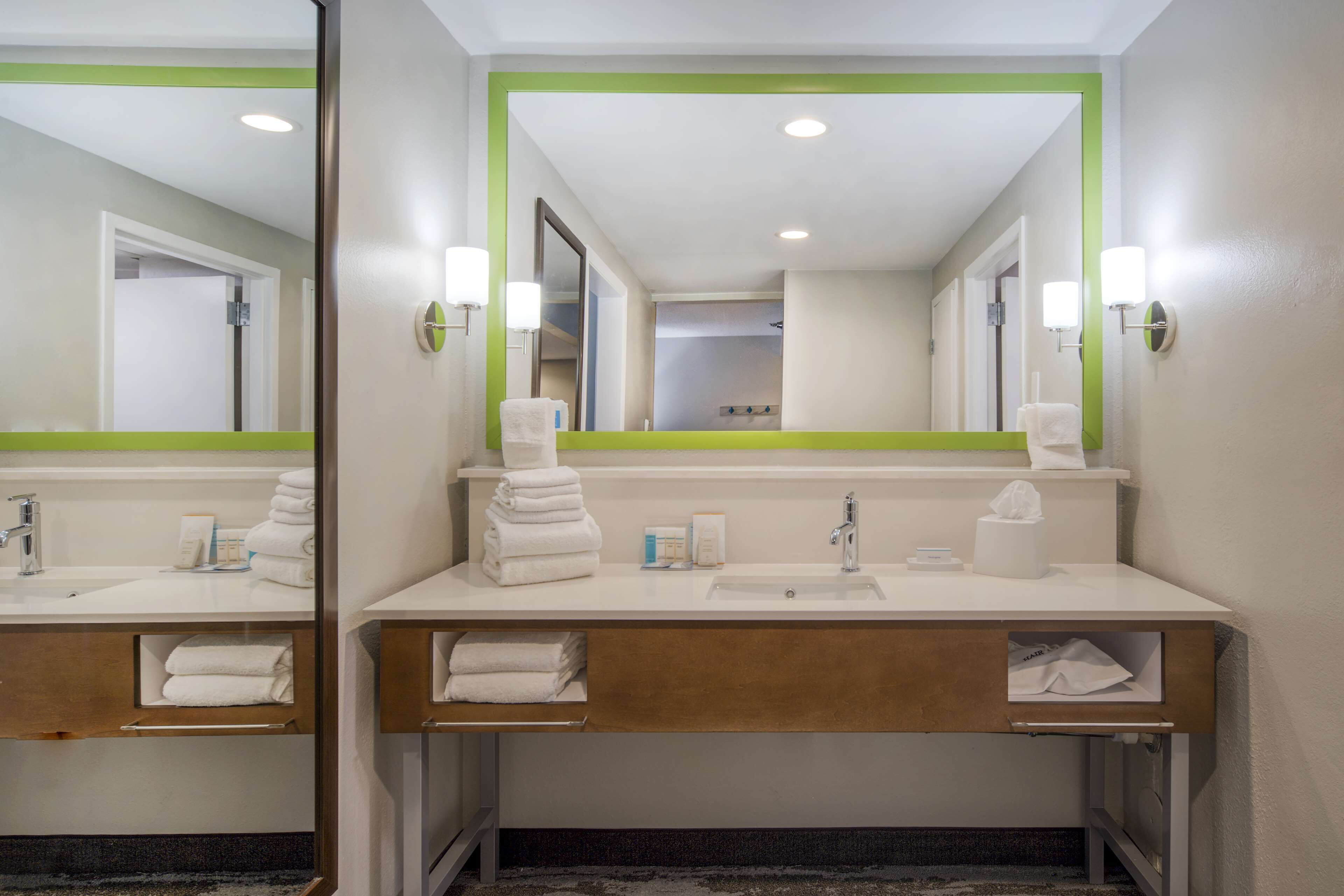 Un baño moderno con un lavamanos blanco, un espejo y un espejo con marco verde. Hay toallas y artículos de aseo en la encimera.