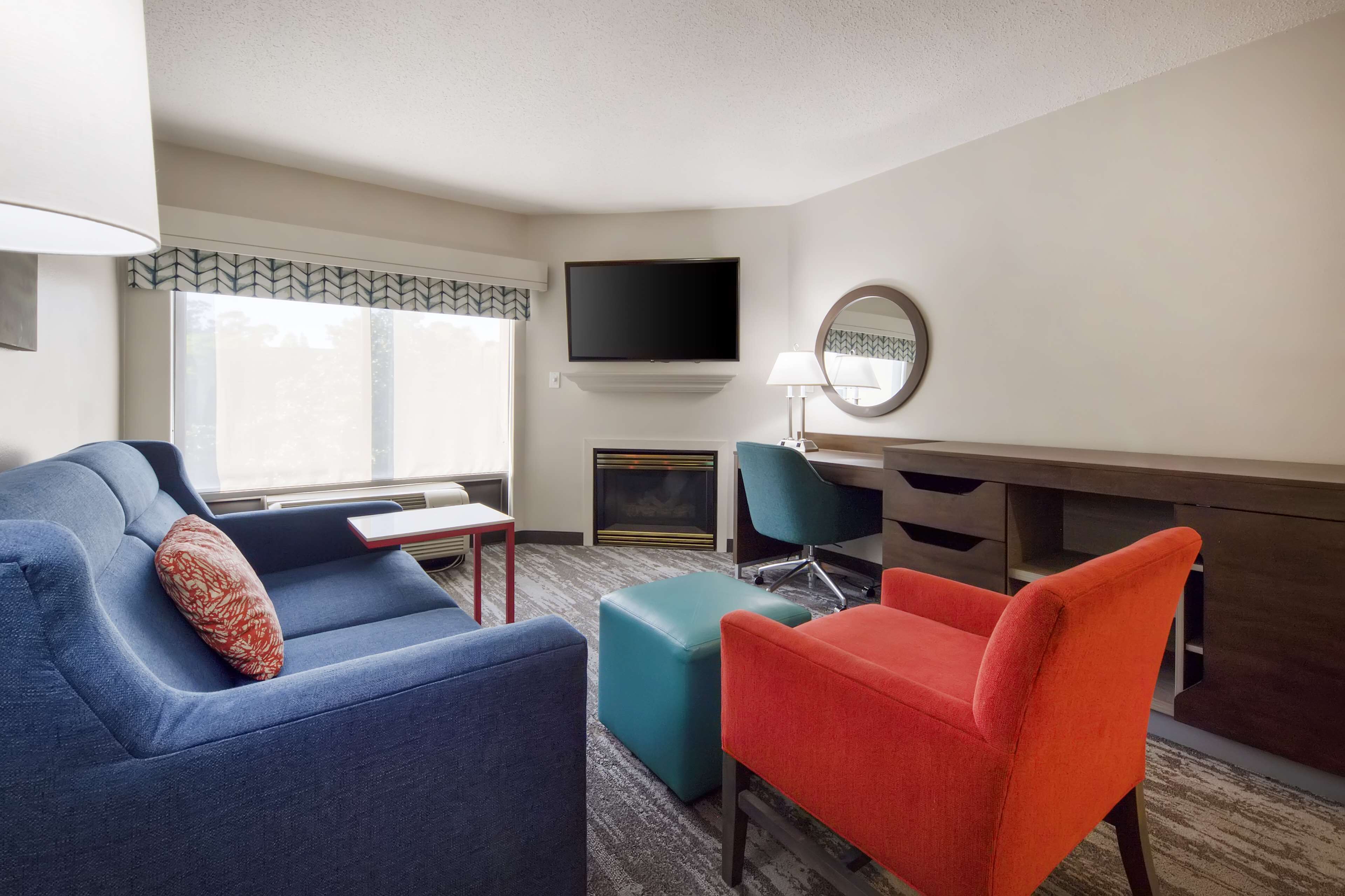 Een moderne hotelkamer met een blauwe bank, een rode fauteuil, een blauwgroene poef, een flatscreen televisie aan de muur, een ronde spiegel en een open haard.