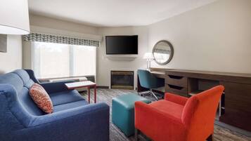 Een moderne hotelkamer met een blauwe bank, een rode fauteuil, een blauwgroene poef, een flatscreen televisie aan de muur, een ronde spiegel en een open haard.