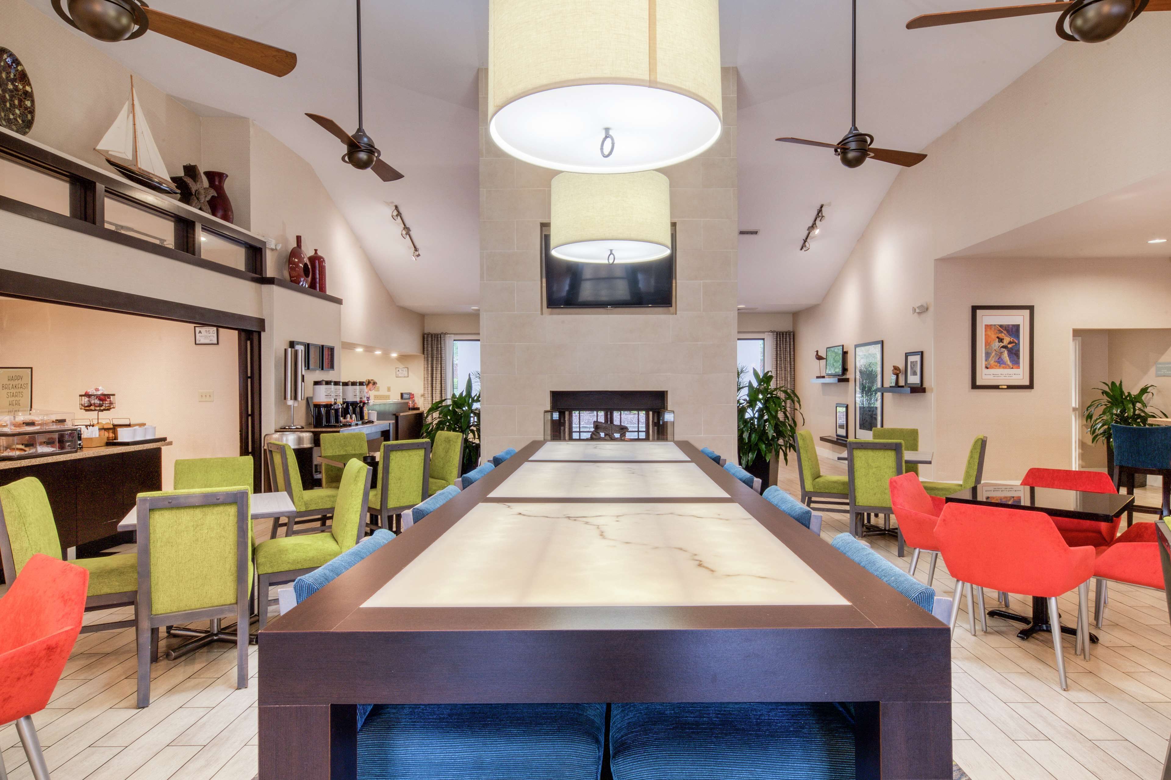 A long communal table with chairs, ceiling fans, and pendant lighting in a modern hotel lobby.