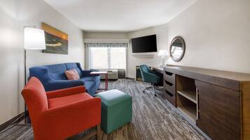 A hotel room with a blue sofa, red armchair, teal ottoman, a desk with a chair, a flat-screen TV, a mirror, and a painting of a sunset.