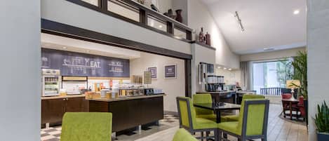 Un moderno vestíbulo de hotel con una barra de café, zona de estar y una escalera que sube a un nivel superior.