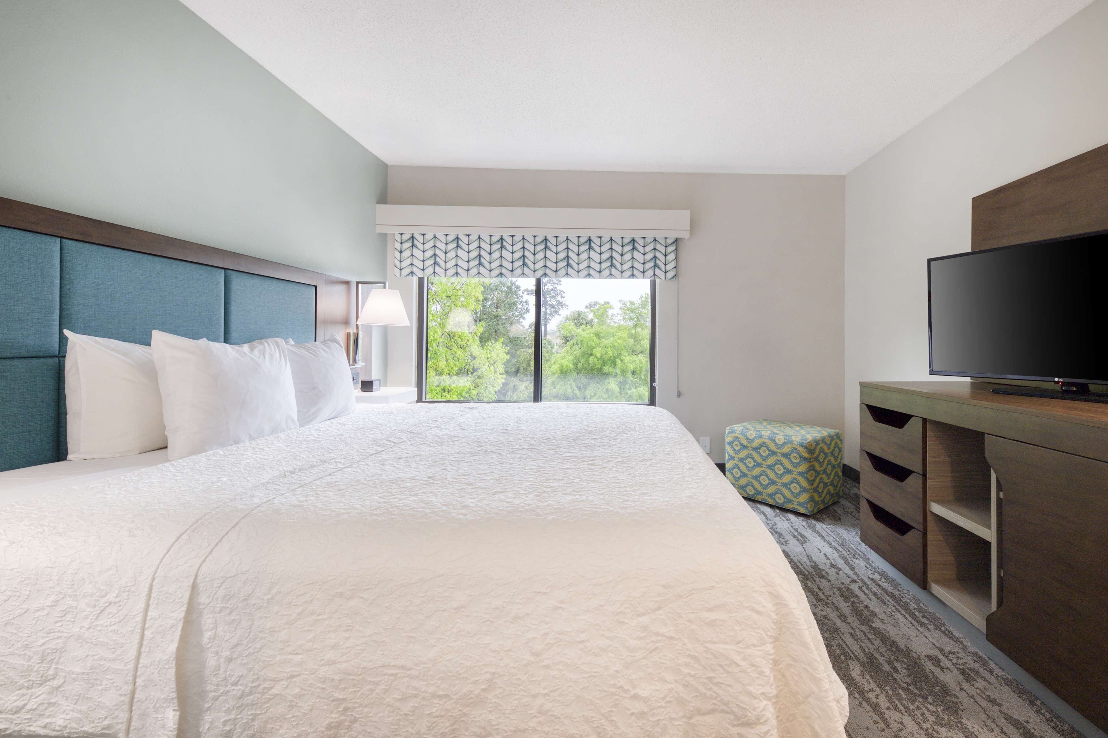 Habitación de hotel con una cama grande, televisor, escritorio y ventana con vista a follaje.