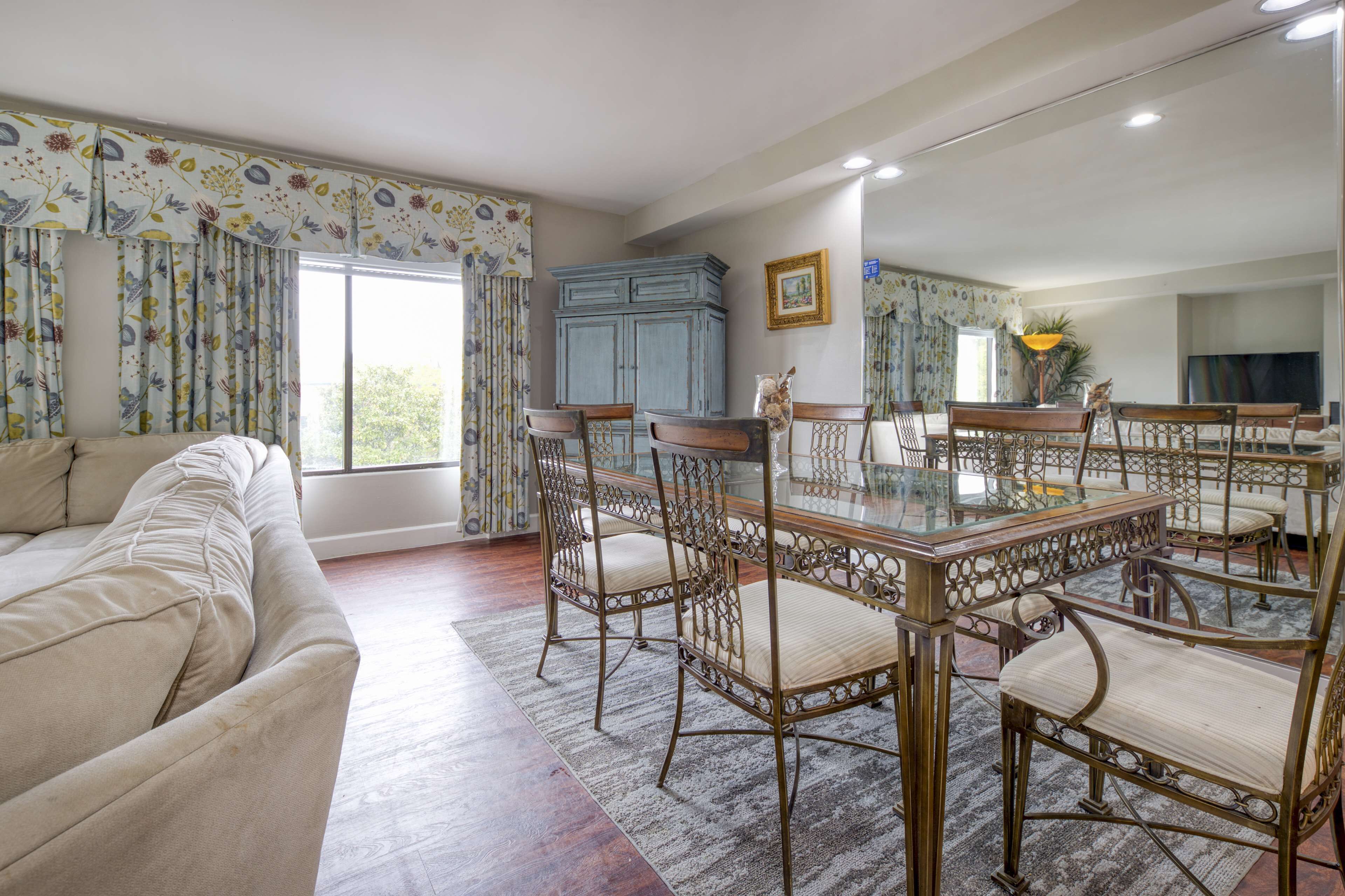 Un comedor con una mesa de vidrio y sillas ornamentadas, un sofá y un espejo grande que refleja la habitación.