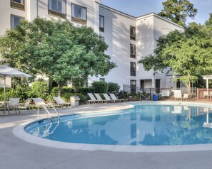 A swimming pool with lounge chairs and umbrellas, surrounded by trees and a building with multiple windows.