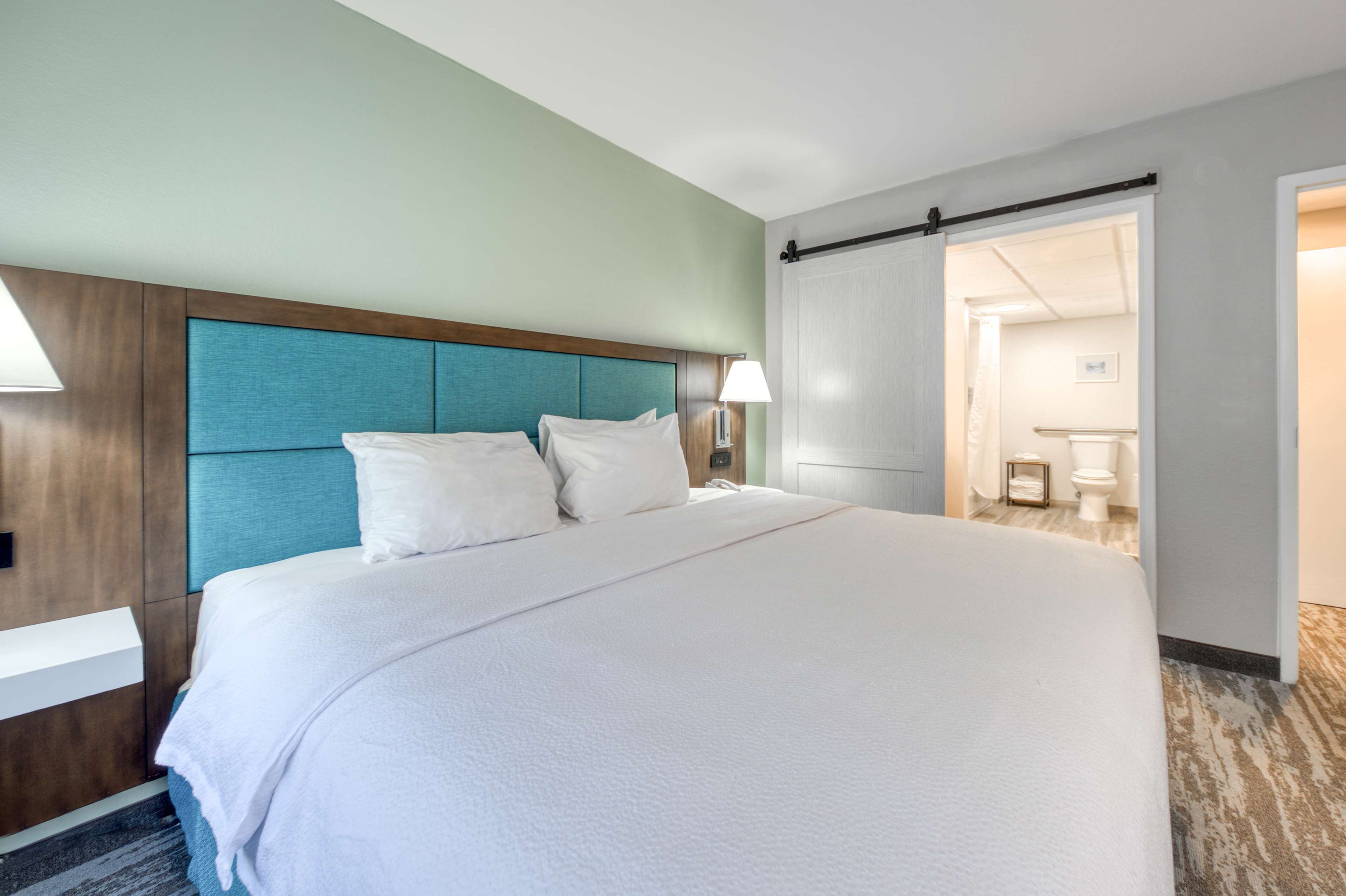 Habitación de hotel con una cama grande, cabecera de madera, mesita de noche y baño visible a través de una puerta abierta.