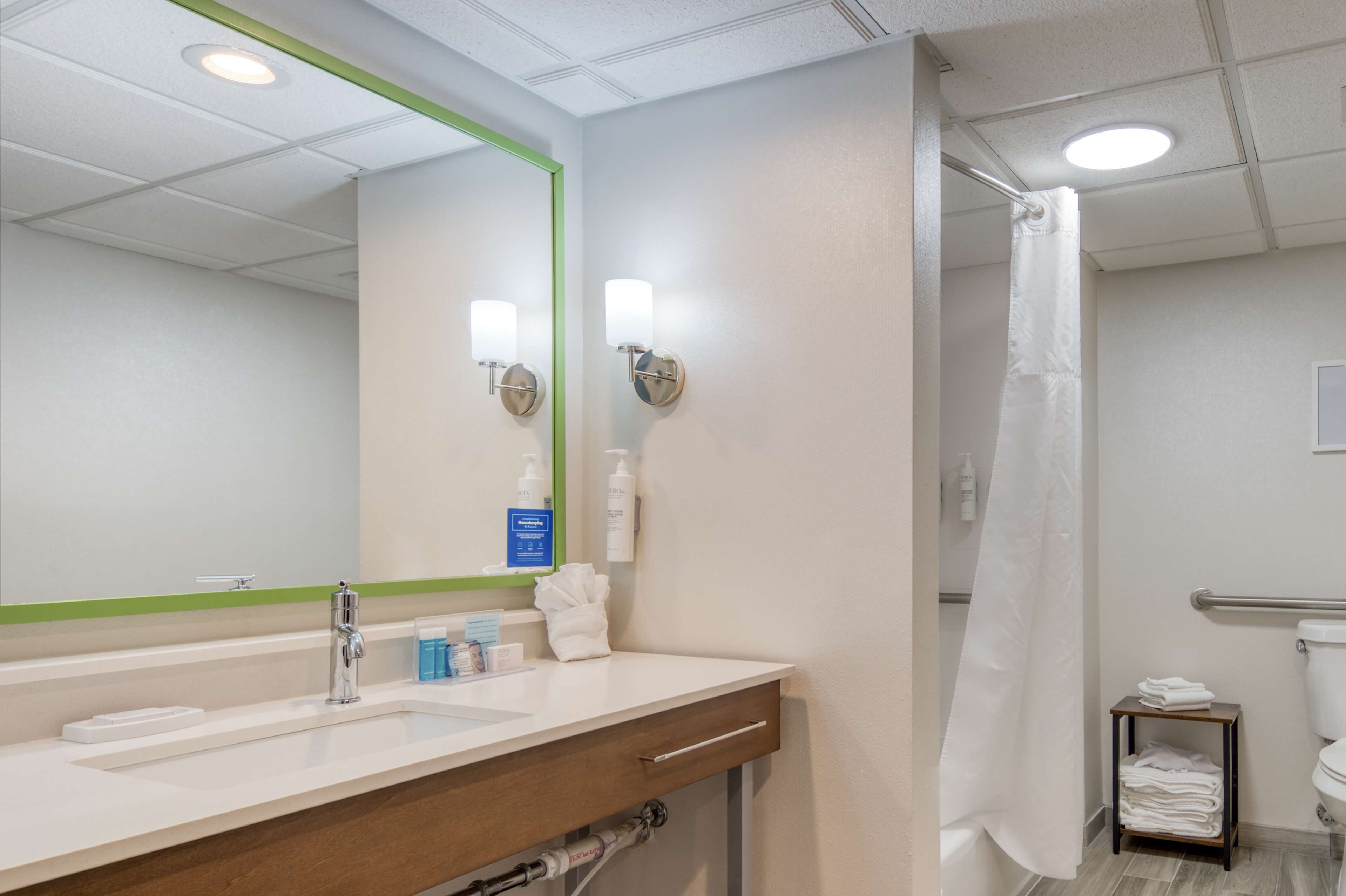 A hotel bathroom with a large mirror, two wall-mounted lights, a shower curtain, a toilet, and a small towel rack.