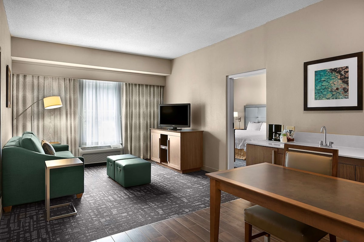 A modern hotel room with a green sofa, a flat-screen TV, a wooden dining table, and a kitchenette.