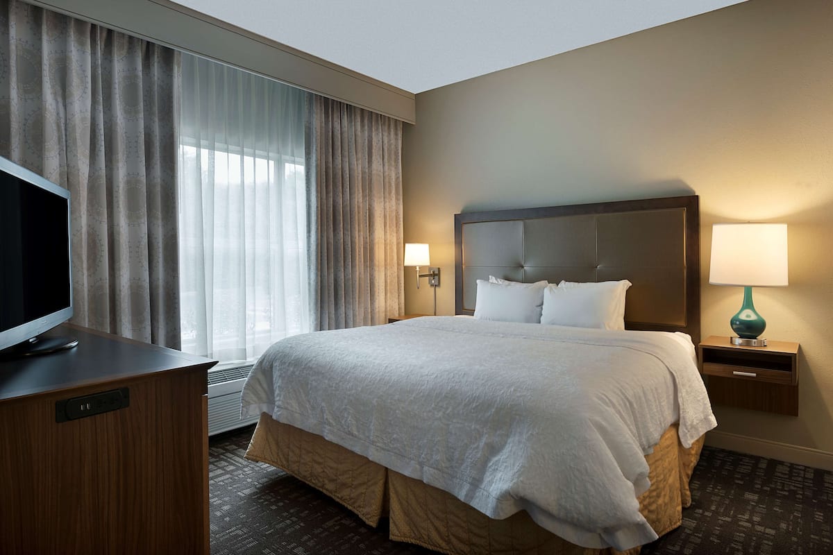 A hotel room with a large bed, a television on a wooden desk, a nightstand with a lamp, and a window with curtains.