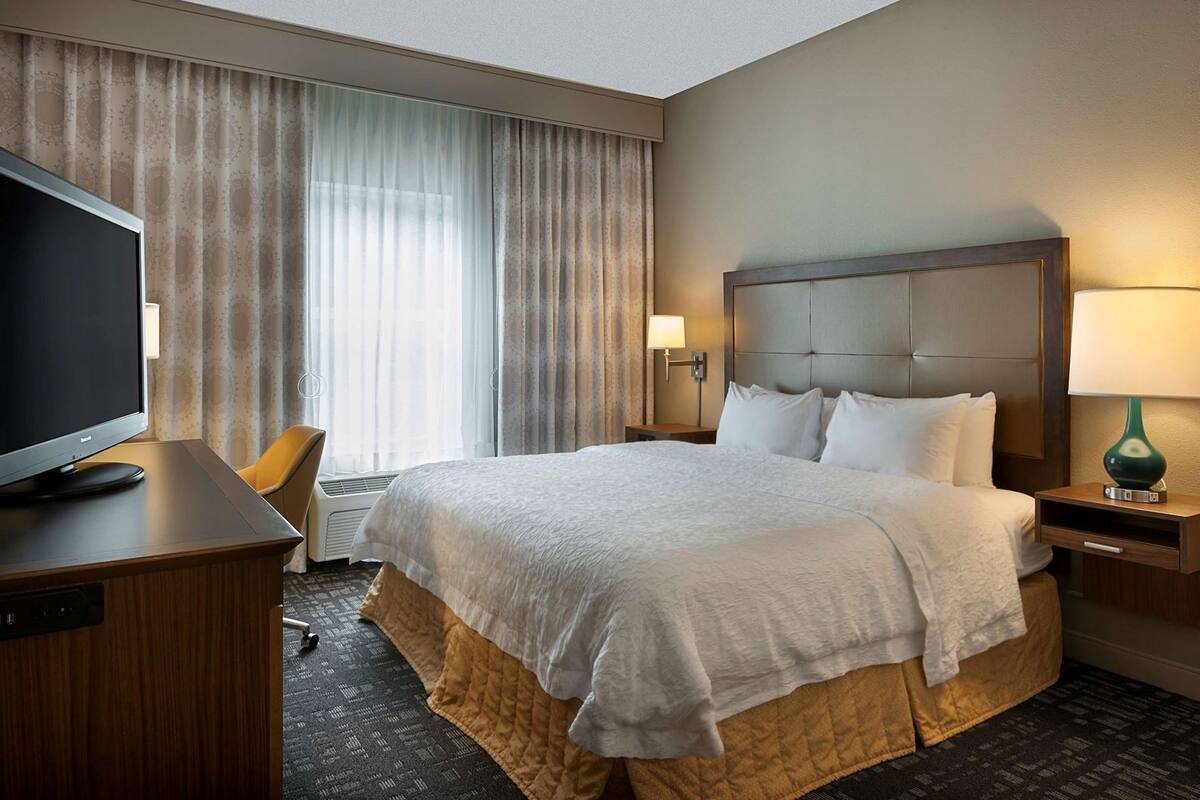 A hotel room with a large bed, a television on a desk, a chair, a lamp, and a window with curtains.
