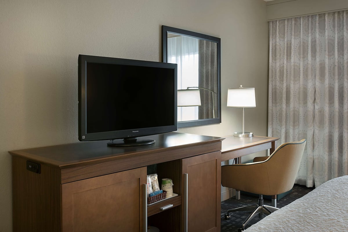 A hotel room with a wooden desk, a television, a mirror, a lamp, and a chair.