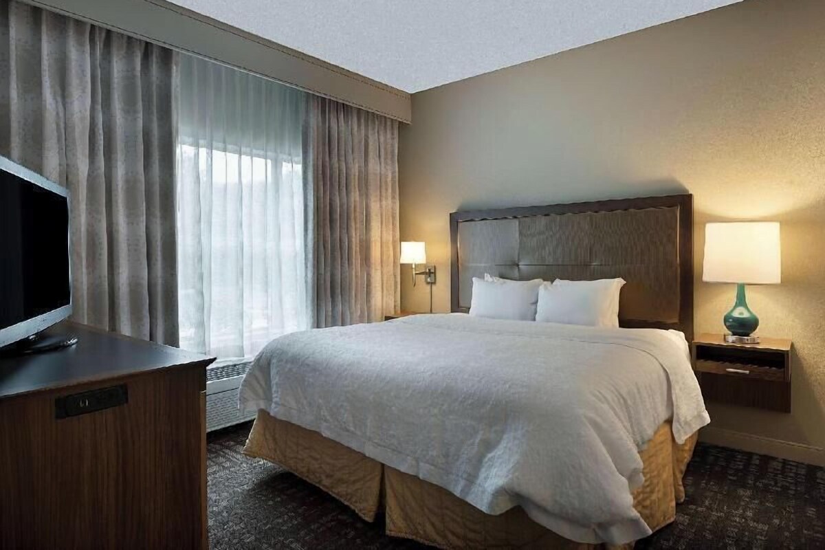 A hotel room with a large bed, a television on a wooden dresser, and a window with curtains.