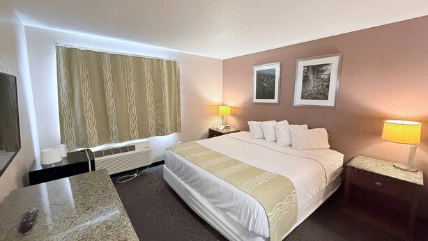 A hotel room with a bed, two bedside lamps, a nightstand, a desk with a telephone, a window with curtains, and two framed pictures on the wall. - Bison Inn & Suites on University (Fargo)