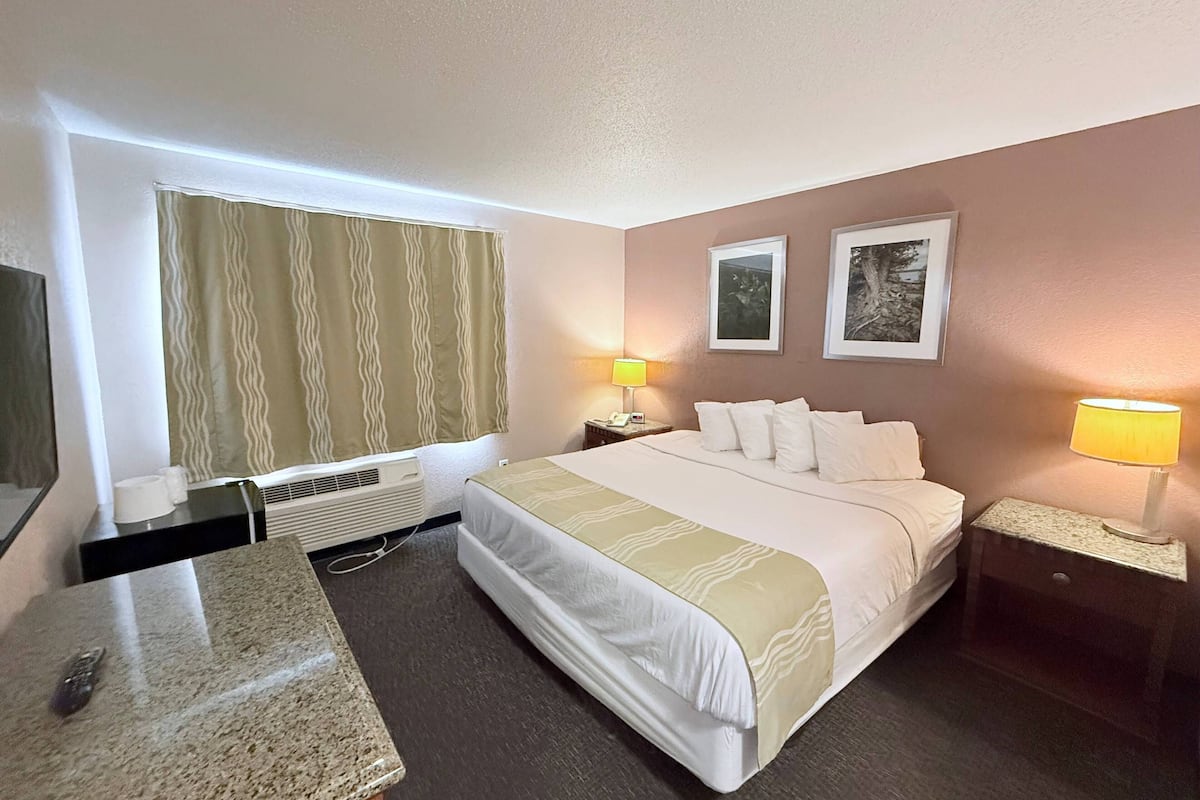 A hotel room with a bed, two bedside lamps, a nightstand, a desk with a telephone, a window with curtains, and two framed pictures on the wall.