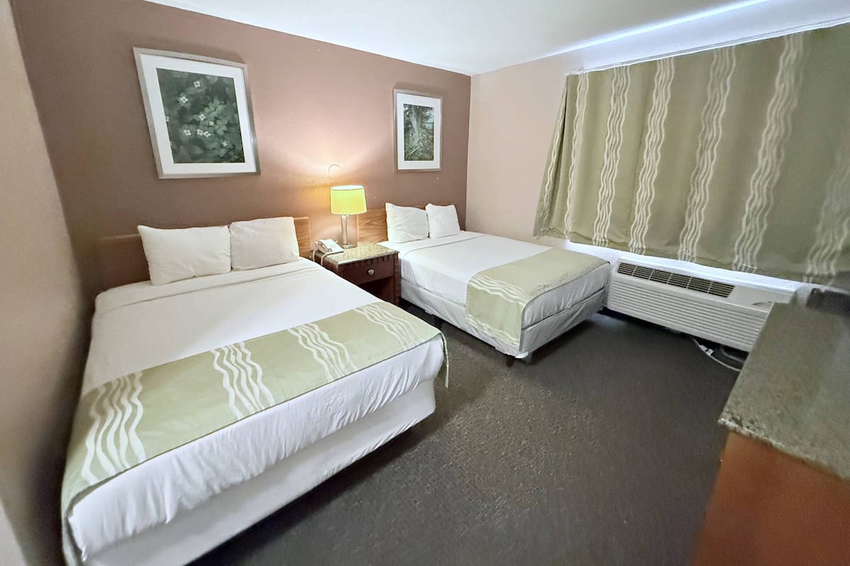A hotel room with two beds, a nightstand with a lamp, a window with curtains, and an air conditioning unit.