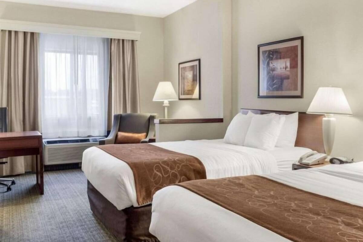 A hotel room with two beds, a desk, a chair, a window with curtains, two wall-mounted lamps, and a framed picture on the wall.