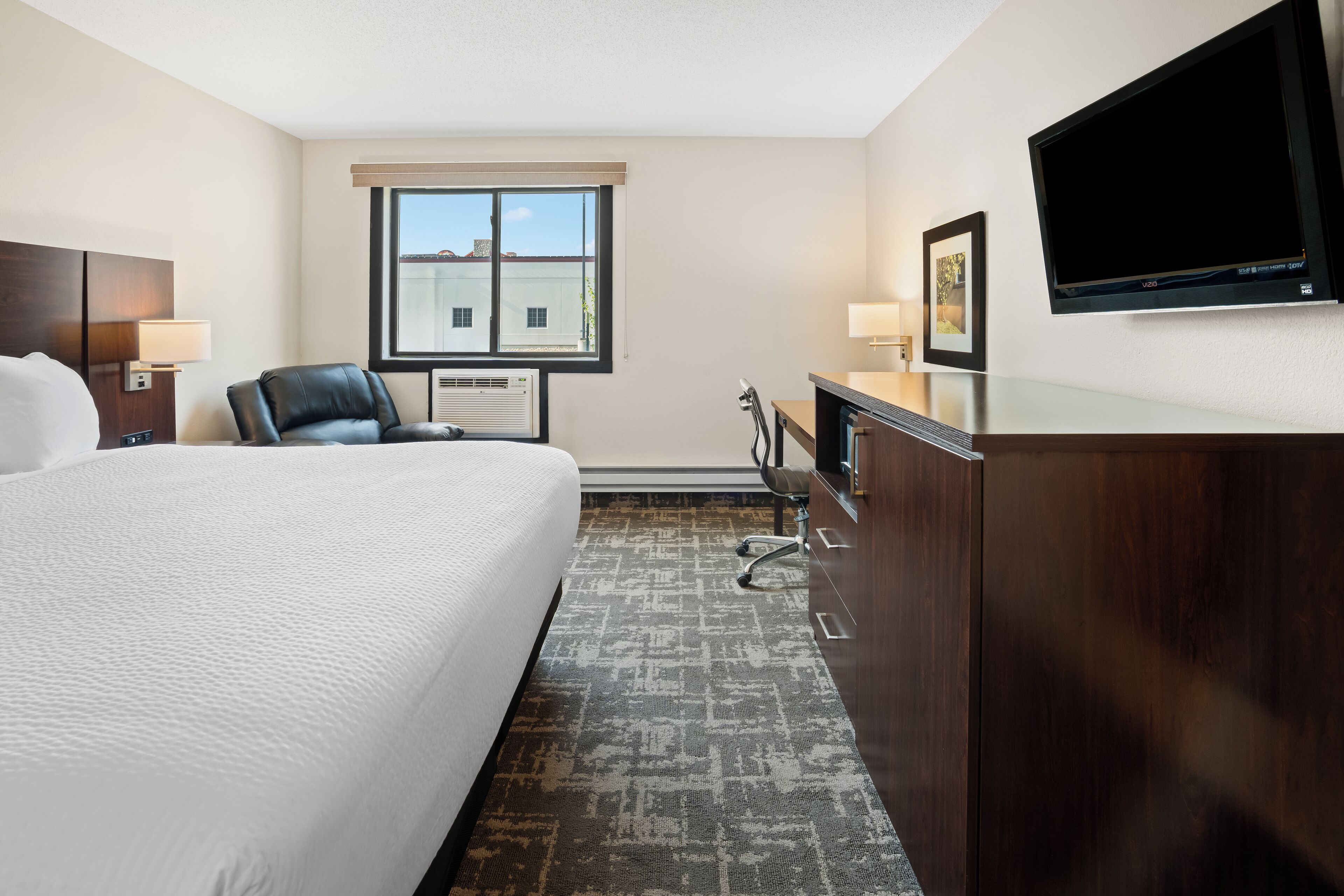 A hotel room with a large bed, a desk with a chair, a TV mounted on the wall, and a window with a view of buildings.