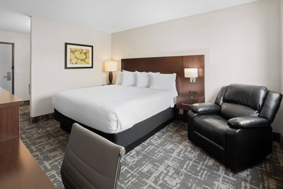 A hotel room with a large bed, a black leather chair, a desk, and a nightstand with a lamp.