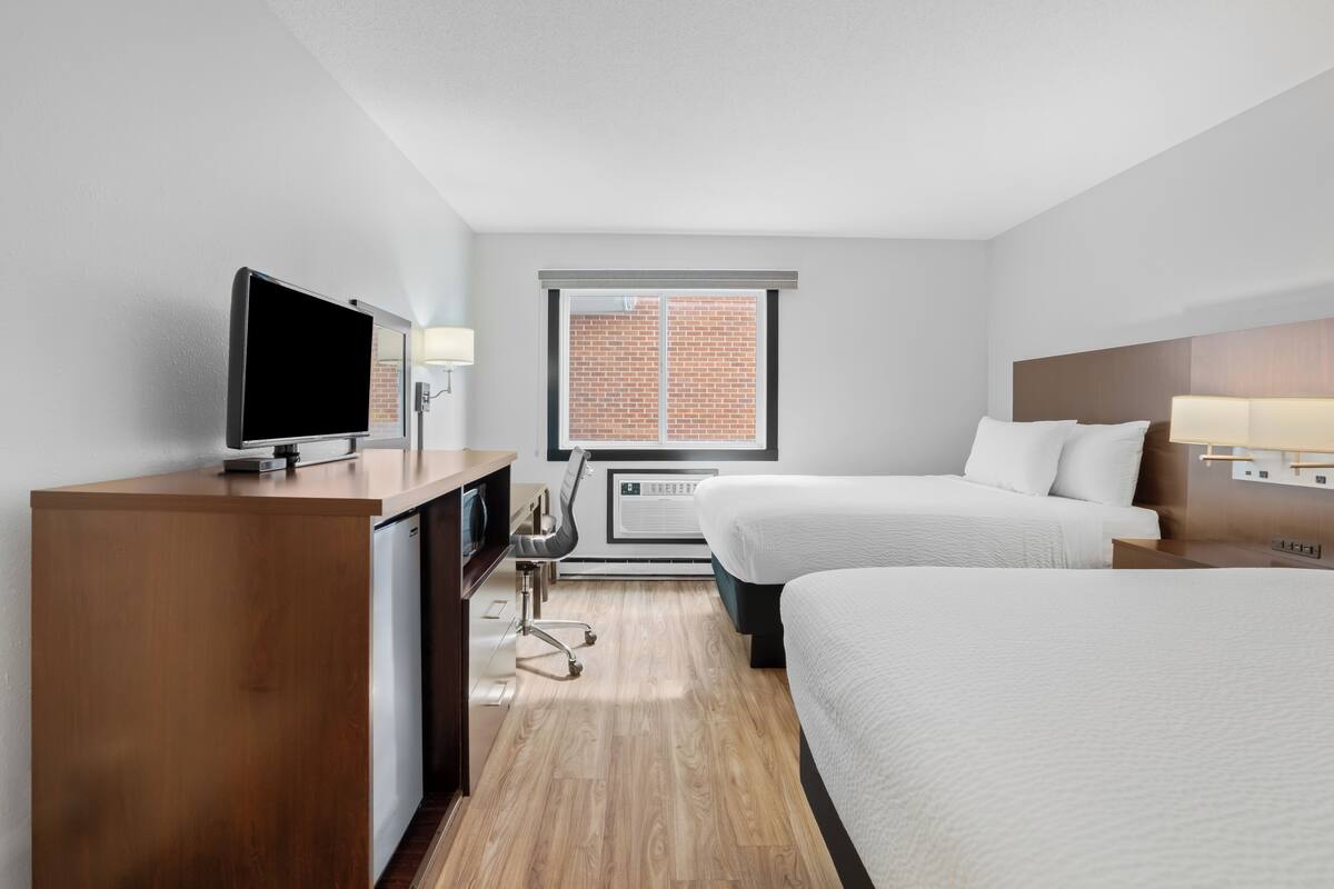 A hotel room with a wooden desk, a television, a window with a view of a brick wall, two beds, and a wall-mounted lamp.