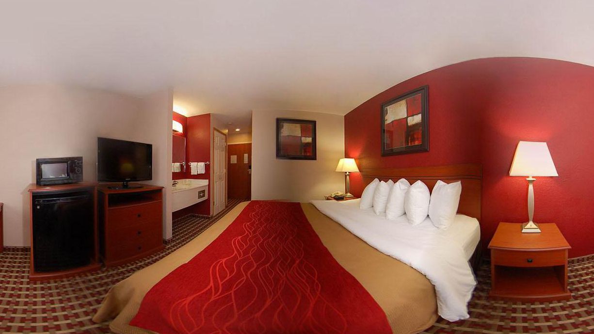 Habitación de hotel con cama, televisor, escritorio y baño visible a través de una puerta abierta.