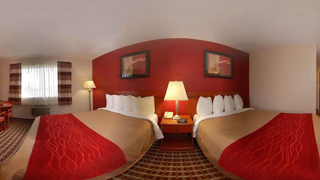 Quarto de hotel com duas camas, uma escrivaninha, uma cadeira, um abajur e duas imagens emolduradas na parede.
