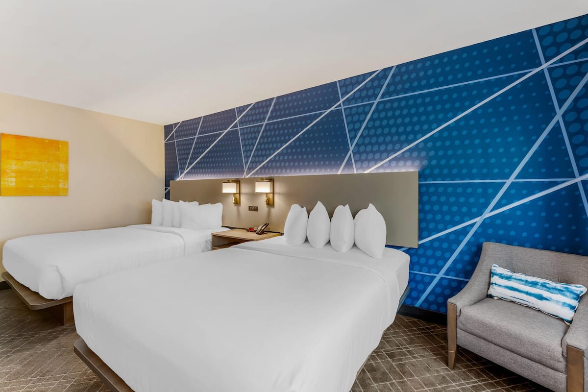 Habitación de hotel con dos camas, un sofá, un escritorio con lámpara y una pared con diseño geométrico.