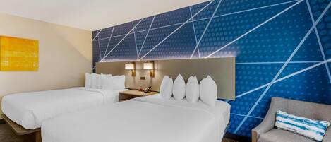 A hotel room with two beds, a sofa, a desk with a lamp, and a wall with a geometric pattern.