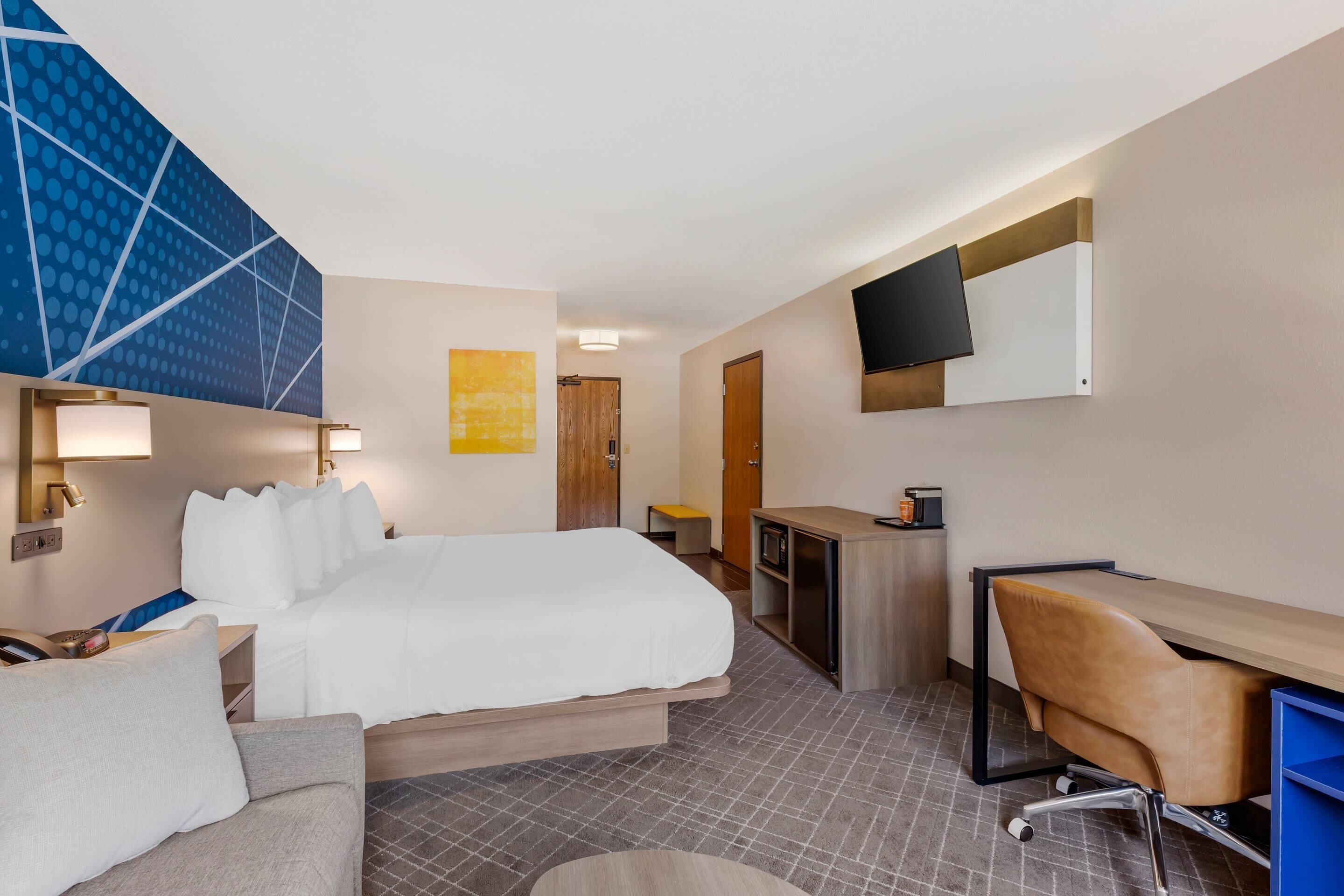 A hotel room with a bed, a desk, a chair, a TV, and a wall-mounted TV.