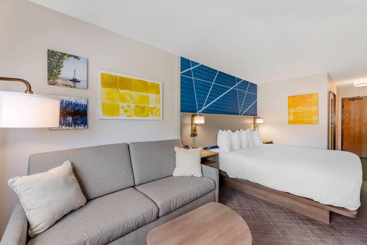 Una habitación de hotel con un sofá gris, una cama, una mesa de madera y obras de arte en las paredes.