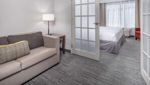 A hotel room with a sofa, a bedside table with a lamp, a large window with curtains, and a bed.