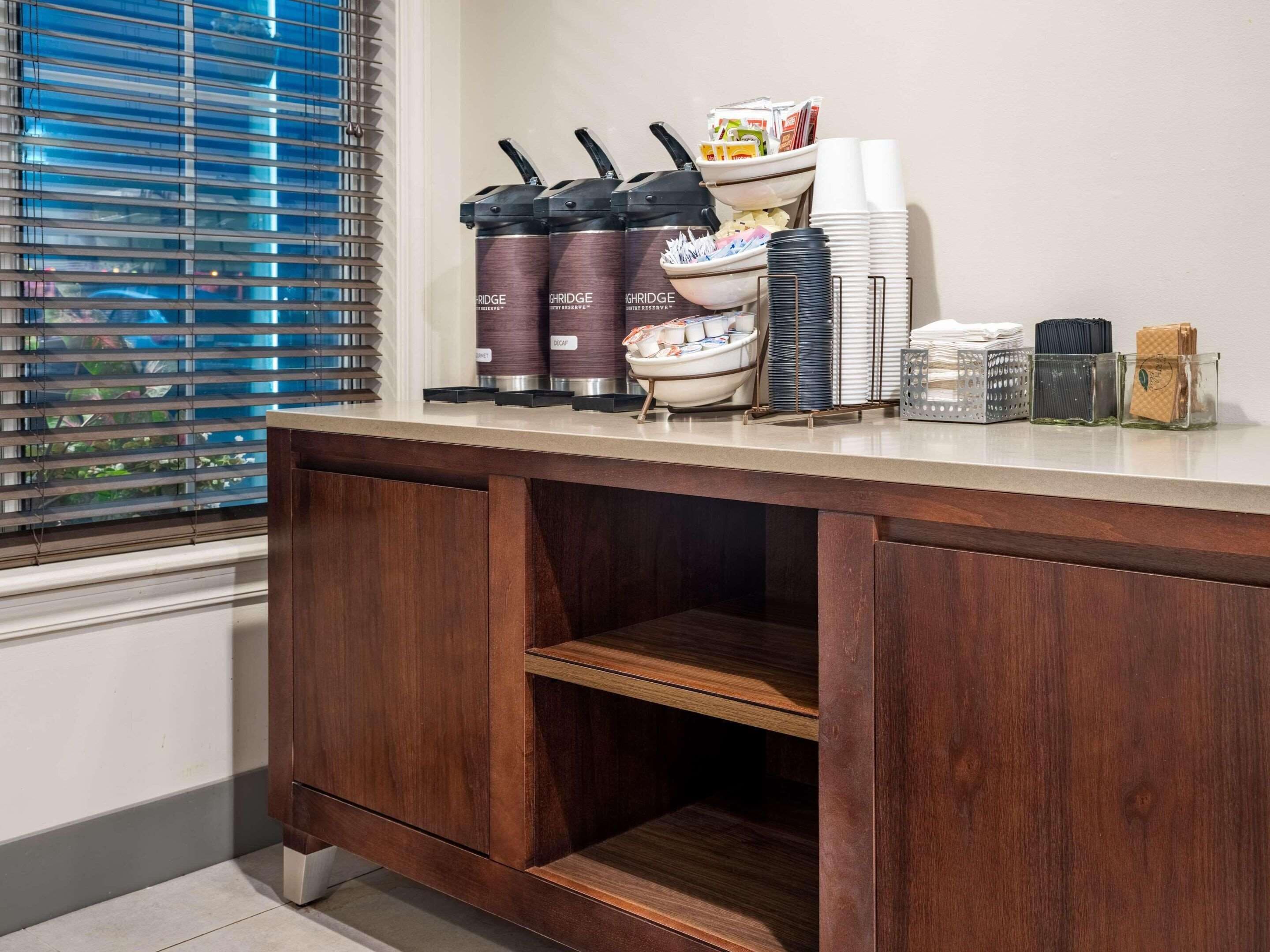 A hotel room with a wooden desk, a window with blinds, and various amenities like coffee makers, bowls, and stacked plates.