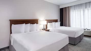 A hotel room with two beds, a nightstand with a lamp, a window with curtains, and an air conditioning unit.