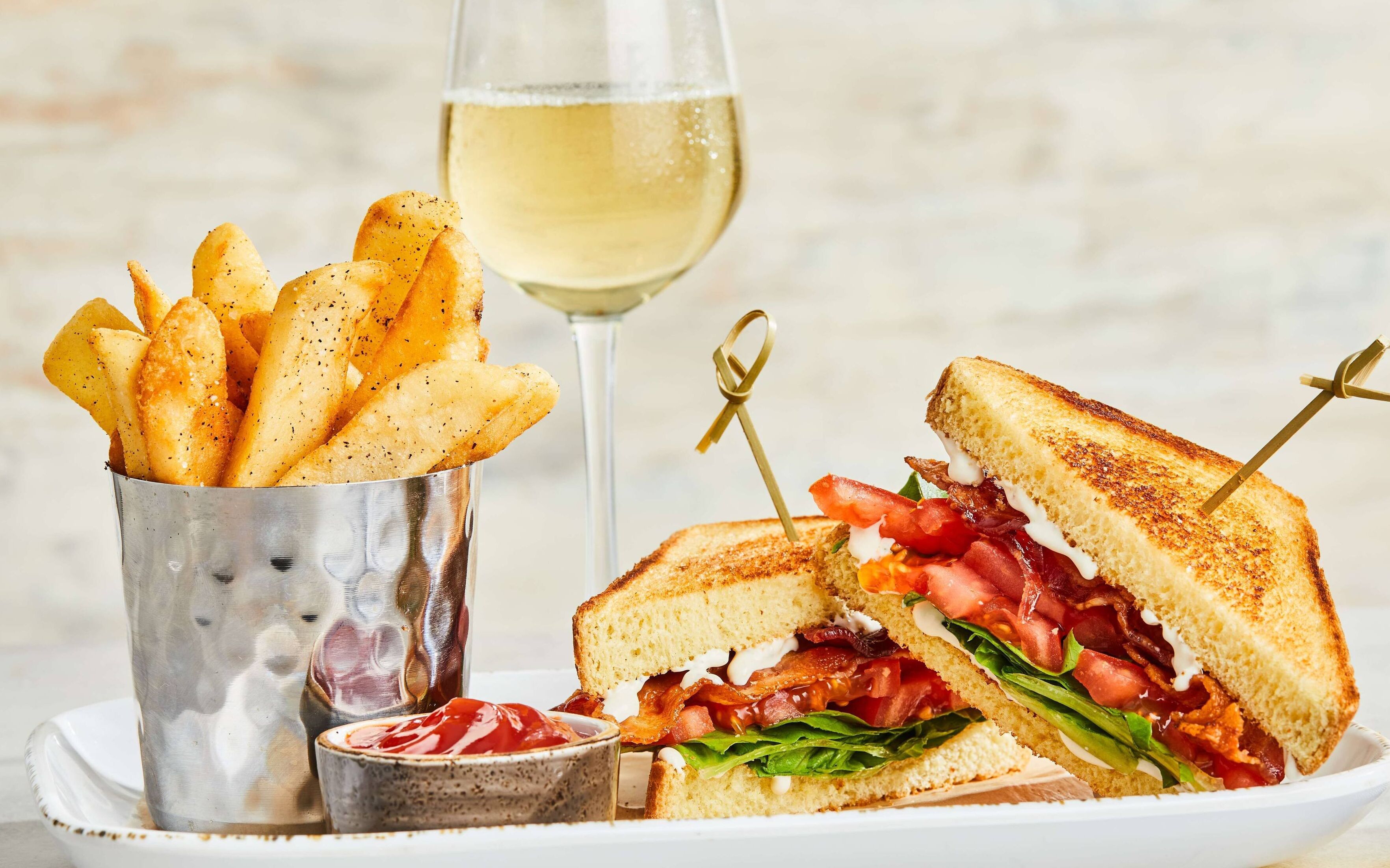 A sandwich with bacon, tomato, and lettuce, a glass of white wine, and a container of fries.