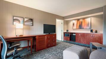 A hotel room with a wooden desk, a television, a kitchenette, and a comfortable seating area.