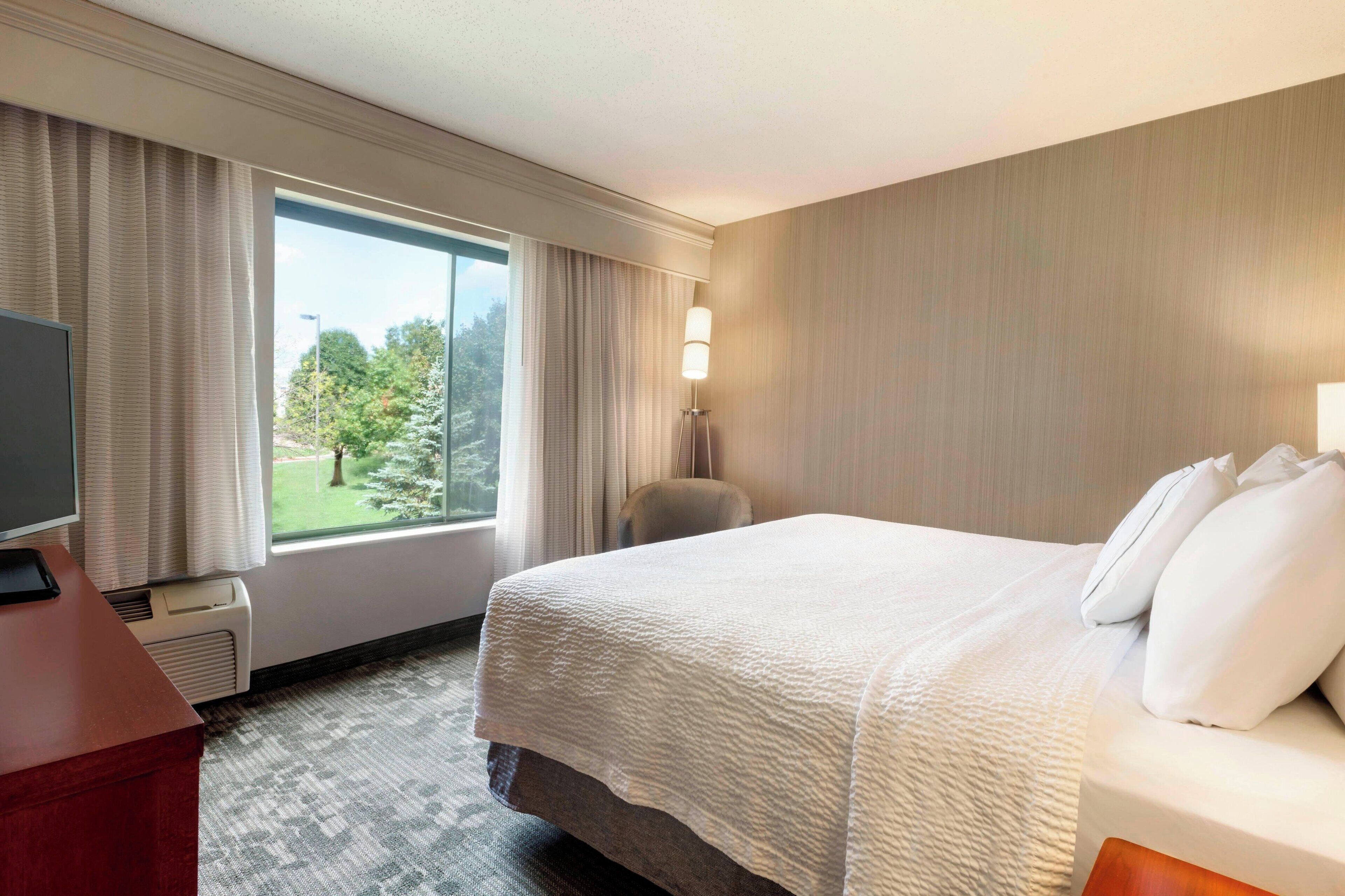 A hotel room with a large bed, a desk with a computer, a chair, and a window with curtains.