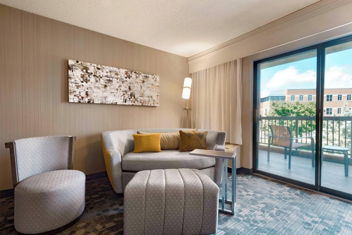 A hotel room with a grey sofa, a matching armchair, a small ottoman, a side table, and a large window offering a view of the outdoors.
