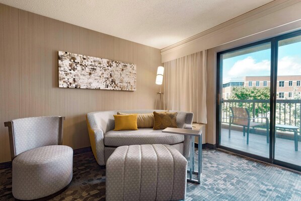 A hotel room with a grey sofa, a matching armchair, a small ottoman, a side table, and a large window offering a view of the outdoors.